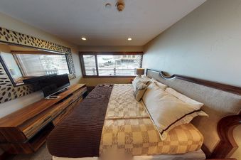 The bedroom at Manor Vail 254 features a king-size bed with a brown and tan checkered comforter, a large window with a view of the mountain, and a wooden dresser with a flat-screen TV.