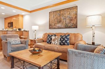 A cozy living room in The Charter G2105 featuring a tan leather sofa, two armchairs, a wooden coffee table, and an adjacent kitchen.