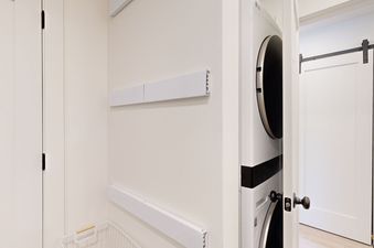 A stacked washer and dryer unit in the laundry area, with white drying racks on the wall and a white wire laundry basket on the floor.