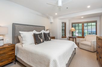 A spacious bedroom features a large bed with a gray upholstered headboard, white bedding, and decorative pillows with deer motifs. Two wooden nightstands with lamps flank the bed, while a cozy armchair and another lamp are by a large window with a view of