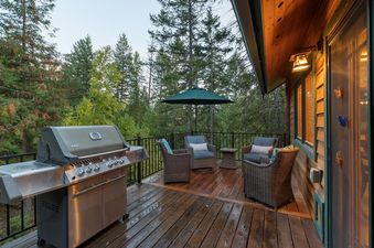 A spacious outdoor deck at Whispering Pines Whitefish with a BBQ grill, seating area, and umbrella, surrounded by pine trees.