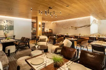 A wide shot of The Sylvan Lodge Two Bedroom Suite's dining area with various seating arrangements including armchairs, banquettes, and tables with place settings, all under a wood-paneled ceiling with modern lighting and a nature-inspired mural.