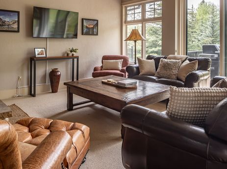 A cozy and rustic living room with leather sofas, a wooden coffee table, and large windows offering views of evergreen trees.