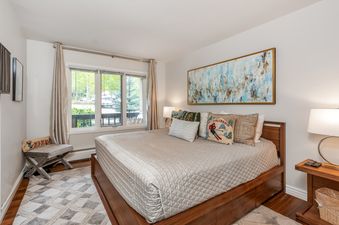 A cozy bedroom in All Seasons 2 Bedroom Platinum Unit B1 with a large bed, decorative pillows, and a window offering a view of lush greenery outside.