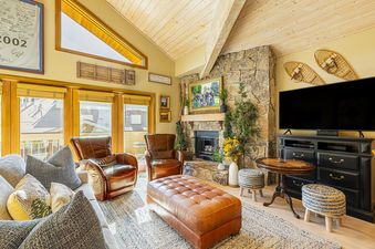 A cozy living room at Royal Plaza 403 featuring a stone fireplace, brown leather furniture, and a large television, illuminated by natural light from ample windows.