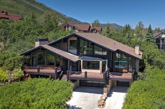 A luxury multi-level house with large windows and decks, nestled among lush green trees on a sunny day.