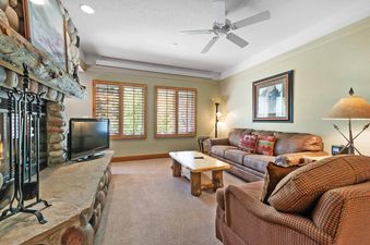The living room of The Charter B1405 features a cozy setup with a leather sofa, plaid armchair, rustic coffee table, and a stone fireplace, all under the soft glow of recessed lighting and a ceiling fan.