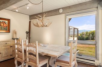 A rustic dining area with a wooden table, chairs, an antler chandelier, and a large window overlooking a scenic mountain landscape.