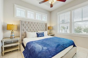 A comfortable king-sized bedroom at Prominence Moby Dick with a tufted headboard, two nightstands with lamps, a blue throw blanket, and windows with blinds.