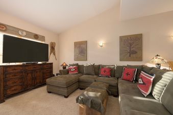 A cozy living room in Waterside West E206 features a large sectional sofa with decorative pillows, a wooden media console, and two framed botanical prints on the wall.