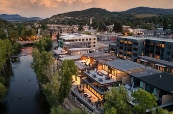 An aerial view of the Yampa River flowing through a town surrounded by green trees and mountains under a sunset sky.