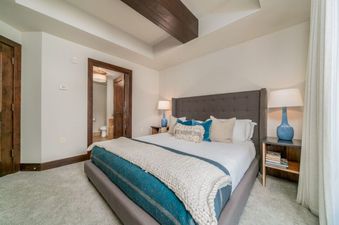 The bedroom in Lumiere 2 Bedroom Suite features a king-size bed with a white duvet, a plush white throw blanket, and a blue throw blanket.  There are blue and tan accent pillows on the bed. The bedside table has a lamp with a blue base and a white shade.