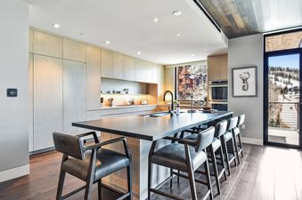 A modern kitchen and dining area in One Empire Pass 303 with light wood cabinets, dark countertops, a large island with five bar stools, and a window offering a view of a snowy mountain landscape.