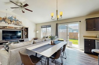 A spacious and inviting living room and dining area, seamlessly blending comfort with functionality, featuring a large, light-colored sofa, a prominent TV, a chic white dining table with a dark blue runner and elegant brown chairs, and a contemporary kitc