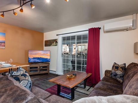 The Resort Plaza 5024 features a cozy living area with an L-shaped sofa, a rustic wooden coffee table, a dining table, and a flat-screen TV, all illuminated by track lighting and natural light from a sliding glass door.