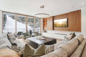 A modern living room with a large sectional sofa, two olive green armchairs, and a dark wooden coffee table, featuring floor-to-ceiling windows overlooking a snow-covered mountain slope with pine trees.