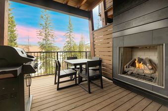 A private balcony at Riverside Reserve features an outdoor fireplace, a dining table with four chairs, and a BBQ grill, all set against a scenic backdrop of pine trees and a colorful sky.