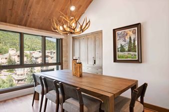 The dining area in Village Hall 13 features a large wooden table, chairs with gray upholstered seats, and an antler chandelier, with a scenic mountain view visible through a large window.