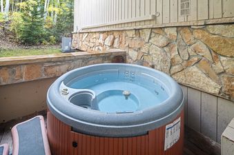 An outdoor hot tub with clear water, set on a wooden deck next to a stone wall and surrounded by trees.