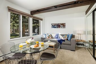 A bright living area in Aspen Alps Apartment 101 features a glass dining table with breakfast, a gray sofa with colorful pillows, and large windows looking out onto trees.
