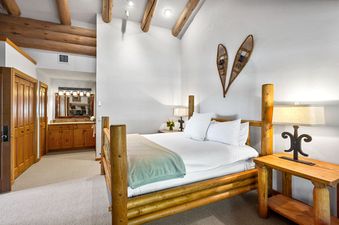 A king-size bed with white linens and a light green blanket is situated in a comfortable bedroom with rustic wooden furniture and decor, a lamp on a nightstand, and decorative snowshoes on the wall.