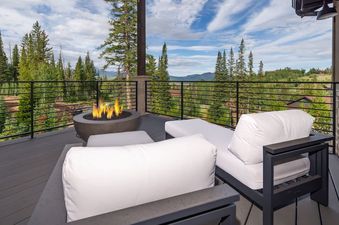 An inviting outdoor deck at Cloud 9 Chalet, featuring a lit fire pit surrounded by modern lounge seating with white cushions, all set against a stunning backdrop of pine forests and distant mountains under a cloudy sky.