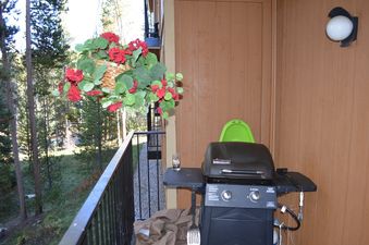 The balcony at Beaver Village 1112 features a black gas grill with a side table, a hanging basket overflowing with red flowers, and a view of pine trees and a gravel path.