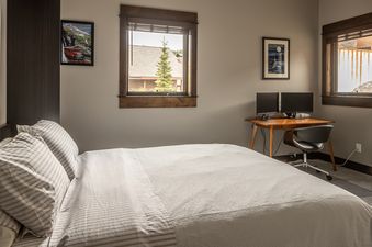 A cozy bedroom in Alpenglow Condo 26D with a bed, a desk with dual monitors, and two windows offering views of the outdoors.