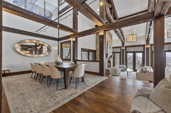 A spacious dining and living area features a large wooden table with chairs, a decorative mirror, and a stone fireplace, all surrounded by large windows looking out onto a snowy landscape.