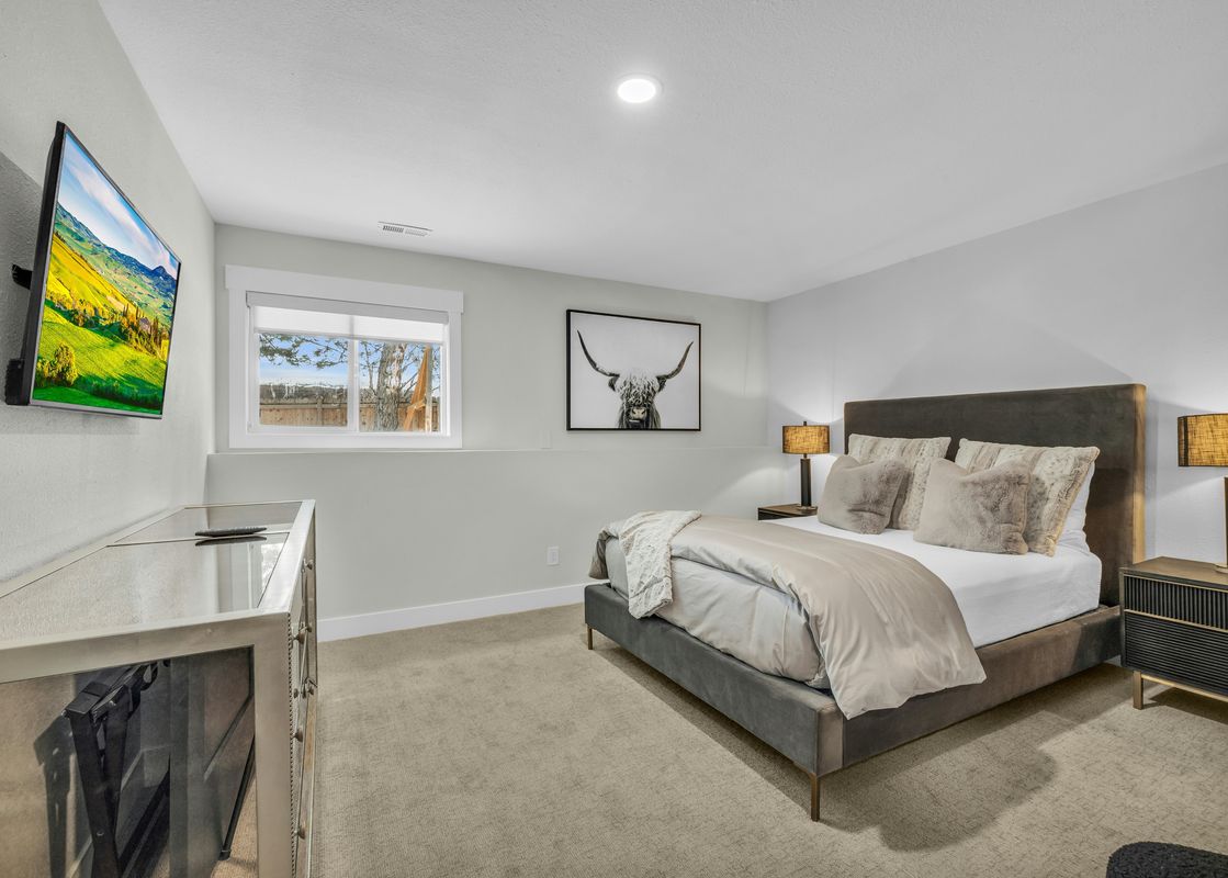 A cozy bedroom with a modern gray bed, two nightstands with lamps, a TV mounted on the wall, and a window with a view of an outdoor fence.