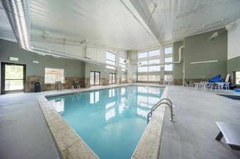 The indoor pool area at Hi Country Haus 309 features a large, inviting pool with clear blue water, surrounded by concrete decking and light-colored tiled edges. The room is well-lit with natural light streaming through numerous large windows and overhead