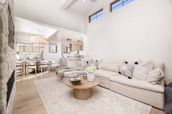A bright and open-concept living room with a large L-shaped sofa, a round coffee table, and a stone fireplace, leading to a dining area and kitchen.