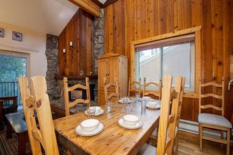 The dining room at Beaver Village 1734 features a rustic wooden table set for a meal, surrounded by matching chairs. A stone fireplace with a wooden mantle and exposed wooden beams add to the rustic charm.