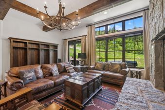 A rustic yet elegant living room with leather sofas, an ornate wooden chest coffee table, an antler chandelier, and large windows showcasing a vibrant green landscape.