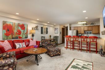 A vibrant living room in Montaneros 211 features a red leather sofa, floral armchair, and a kitchen island with red bar stools, all under warm recessed lighting.