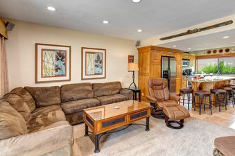 A cozy brown sectional sofa, coffee table, and leather recliner in the living room, with a modern kitchen featuring an island and bar stools in the background.