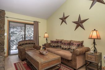 A cozy living room at Beaver Village 232 features a comfortable brown sofa and armchair, a rustic wooden coffee table, and large windows revealing a snowy forest.