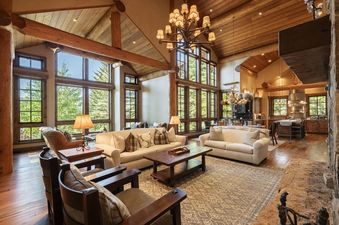 The Deer Valley Oak Lodge features a vast living area with high ceilings, exposed wooden beams, and large windows that offer views of lush greenery.