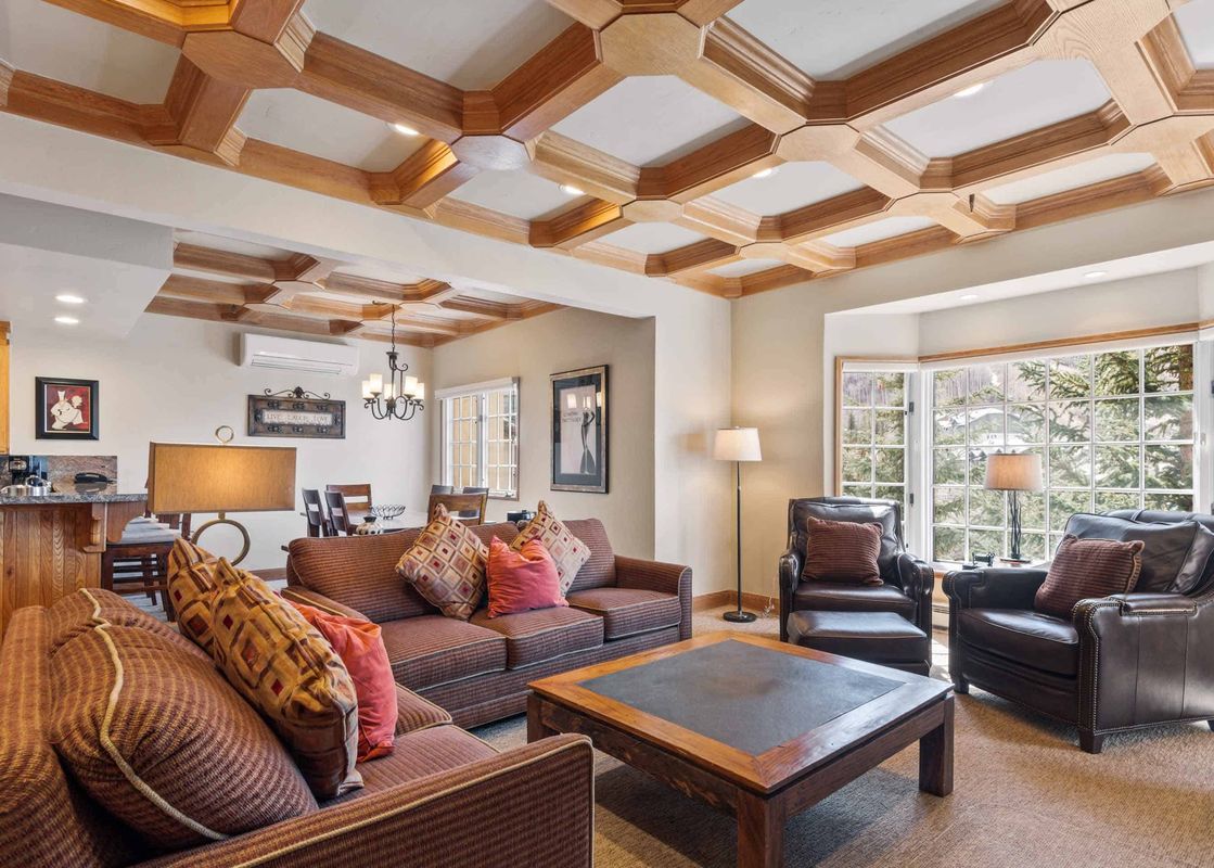 The living room of The Charter F3205 features brown sofas, dark leather armchairs, and a wooden coffee table under a coffered ceiling, with large windows overlooking snow-covered trees.