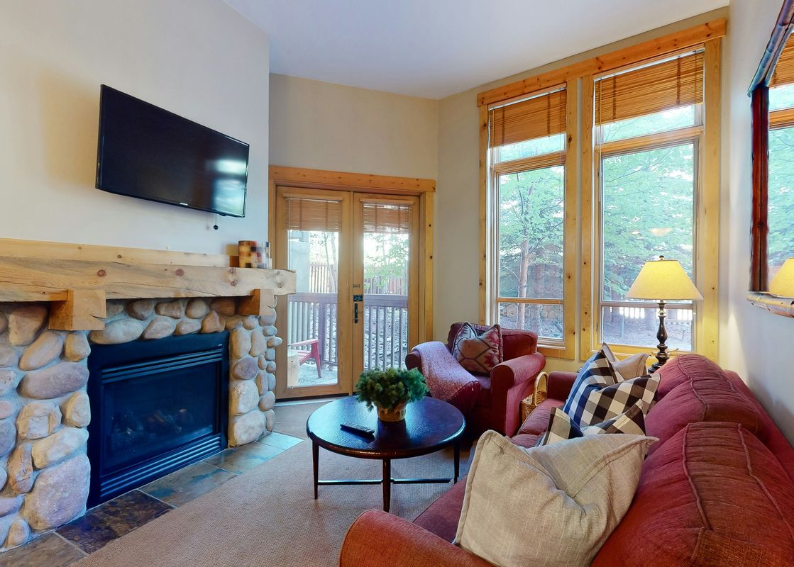 The living area at Taylors Crossing 109 features a comfortable red sofa and armchair, a fireplace with a stone facade, and large windows that offer views of the lush green trees outside.