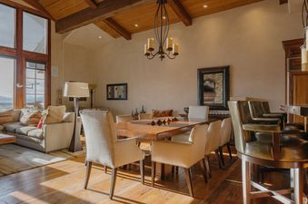 A wide view of the dining and living area at Lynx Lookout at See Forever, showcasing a wooden dining table with upholstered chairs, a comfortable sofa, and large windows with an exterior view.