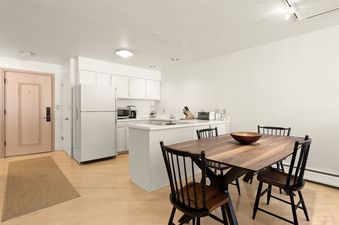 A bright and open-plan kitchen and dining area in Southpoint 2D, featuring white appliances, light hardwood floors, and a dark wood dining table with black chairs.