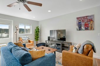 A cozy living room featuring a blue sofa, brown leather armchairs, a wooden coffee table, a flat-screen TV on a media console, and large windows with a view of a snowy landscape.