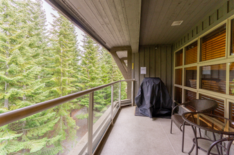 A private balcony at Aspens on Blackcomb Unit 427, featuring a covered BBQ grill and outdoor seating, with a lush forest view.