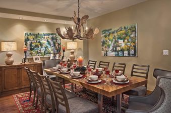 A rustic dining room with an antler chandelier, a long wooden table set with red placemats and candles, and two vibrant paintings on the walls.