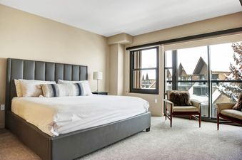 A cozy bedroom with a large bed, two armchairs by a window with a snowy view, and a neutral color palette.