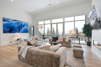 A spacious and modern living room with large windows overlooking a snow-covered forest, featuring light-colored sofas, a gas fireplace, and a vibrant blue abstract painting.