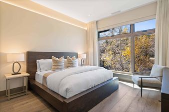 A modern bedroom with a large bed, two nightstands with lamps, and a window overlooking autumn trees.