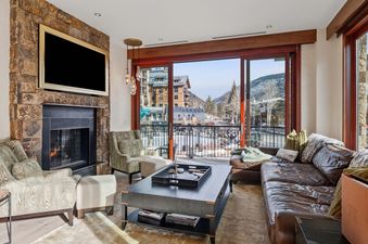 A cozy living room in Solaris 2A South features a stone fireplace, a large TV, and comfortable seating, with an expansive view of a mountain village and snowy peaks from the balcony.