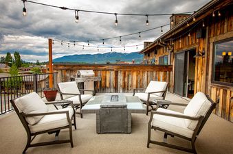 An outdoor terrace at Cana Loft 302 features comfortable seating, a fire pit table, a barbecue grill, and string lights, all set against a mountain backdrop under a dramatic sky.